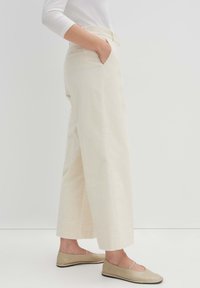 Weite, cremefarbene Cordhosen mit glatter Textur und Fronttaschen, kombiniert mit hellbeigen, flachen Slippers.