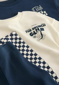 Dwie złożone bluzy, jedna biała z tekstem "The Brunch Club" i kreskówkowym awokado, druga granatowa z postacią grzanki, zestawione z niebieskimi spodenkami w kratę.