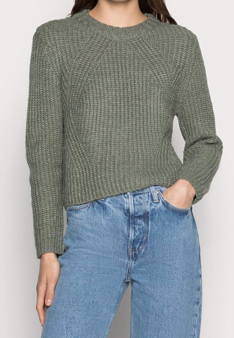 Femme portant un pull en maille texturée vert olive à manches longues et un jean taille haute bleu clair, une main dans la poche.