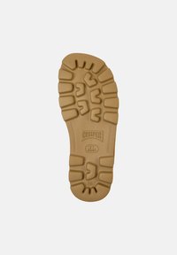 Gummisulor i tan färg, med texturerade mönster och ett stämplat logotyp. Släta kanter med definierade dämpningsområden.
