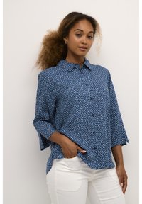 Chemise bleu à manches boutonnées avec un motif floral, coupe ample et manches 3/4. Fabriquée en tissu léger, elle présente un col pointu et des fentes sur les côtés.
