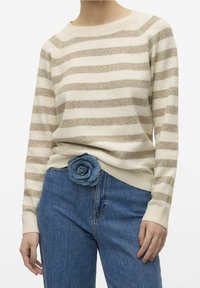 Beige- und cremefarbener gestreifter Pullover mit Rundhalsausschnitt, getragen über blauen Jeans. Ein Denim-Blumen-Detail ist am Bund angebracht.