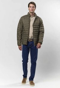 Uomo che indossa una giacca trapuntata verde oliva sopra una camicia beige, jeans blu, cintura marrone e sneakers marroni, in piedi su uno sfondo bianco uniforme.