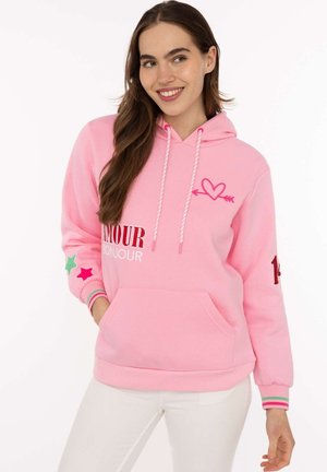 Frau trägt pinken Hoodie mit Herz- und Pfeilgrafik, "AMOUR BONJOUR" Schriftzug, Stern-Patches am Ärmel und weiße Hose, lächelt leicht.