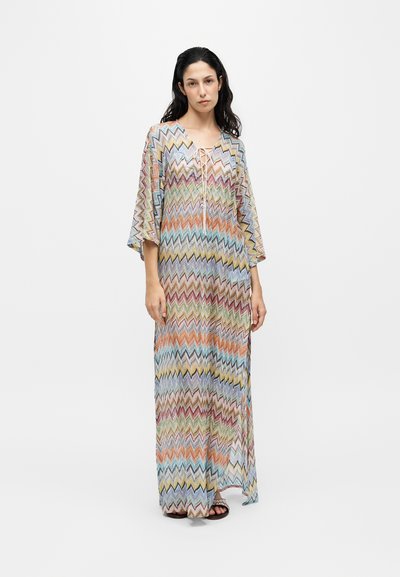 Femme aux longs cheveux foncés portant une longue robe multicolore à motif zigzag avec de larges manches et un col lacé, debout sur un fond uni.