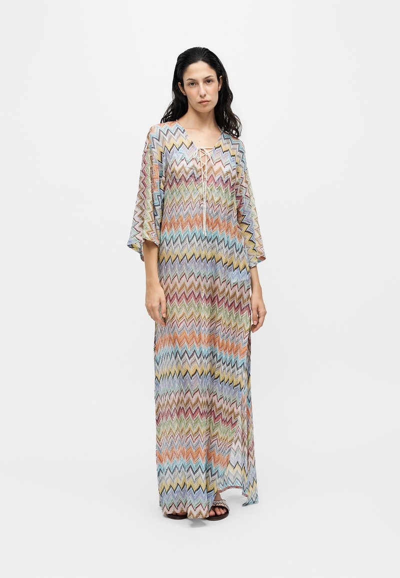 Femme aux longs cheveux foncés portant une longue robe multicolore à motif zigzag avec de larges manches et un col lacé, debout sur un fond uni.