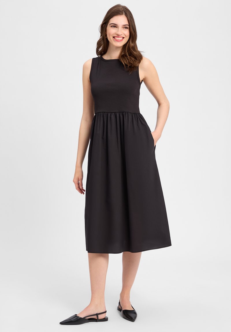 Schwarzes Kleid mit einem taillierten Tank-Oberteil, gerafftem Bund und fließendem Rock. Der Stoff sieht glatt aus, mit seitlichen Taschen und einem knielangen Saum.