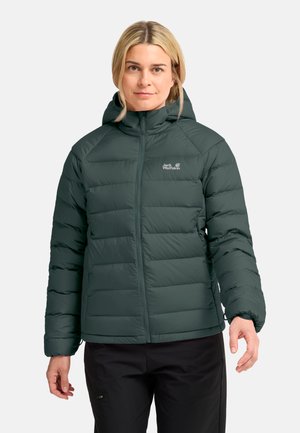 Vrouw met een donkergroene gewatteerde jas met capuchon en een rits aan de voorkant en zwarte broek, staand tegen een effen witte achtergrond.