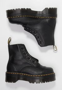 Une paire de bottes de combat en cuir noir avec des semelles épaisses à rainures, des coutures jaunes, des œillets en métal et des languettes de tirage sur un fond blanc.