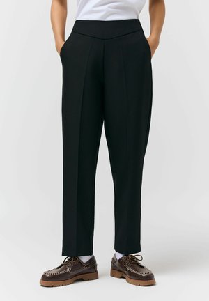 Osoba ubrana w czarne spodnie na miarę z przednimi kantami, białe skarpetki, brązowe mokasyny sznurowane i biała koszula, z rękami w kieszeniach.