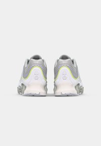 Sive in bele športne čevlje z teksturirano zgornjim delom, ki imajo neonsko rumene detajle in oblikovano podplat za dodatno blaženje.