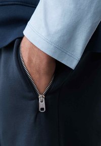 Tessuto blu navy con un dettaglio della cerniera argentata su una tasca, completato da una manica azzurro chiaro con cuciture visibili. Ingrandimento della zona della tasca.
