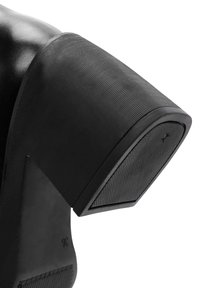 Chaussure noire à talons hauts avec un talon en caoutchouc texturé. Le talon a une forme rectangulaire et un bord légèrement courbé pour plus de stabilité.