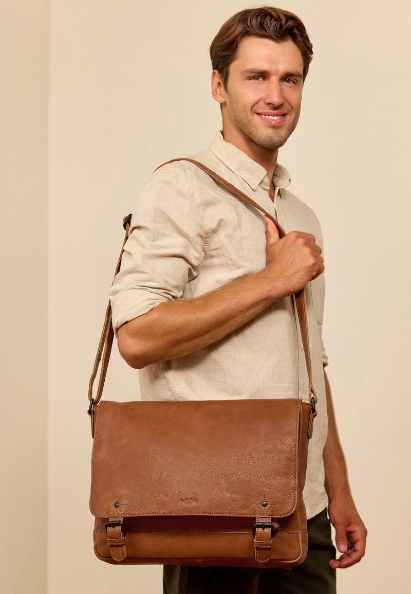 Braune Leder-Umhängetasche mit verstellbarem Gurt, ausgestattet mit einem Klappenverschluss und dekorativen Schnallen. Glatte Textur und minimalistisches Design.