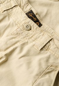 Pantalones cortos de algodón beige con una textura suave, que cuentan con un cierre delantero de botón y una etiqueta cosida con la marca "Superdry" en la cinturilla.