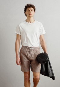T-shirt blanc à manches courtes, shorts marron avec cordon de serrage, et veste en cuir noire tenue dans la main droite. Textures lisses, coupe décontractée, couleurs neutres.