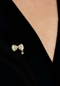 Gold-tone brooch designed as a bow, featuring clear crystal accents and a small charm, pinned on a dark fabric surface.
