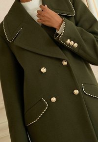 Maslinasto zeleni kaput na dvostruko kopčanje s zlatnim dugmadima, velikim ovratnikom i crno-bijelim detaljima na džepovima i rukavima.