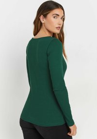 Pitkähihaine ribattu paita tummanvihreänä, jossa on pyöreä kaula-aukko ja istuva siluetti, joka sujuvasti muuttuu kevyesti leveneväksi helmaks.