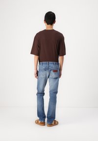 Camiseta marrón de manga corta, jeans de mezclilla azul claro y sandalias beige. Los jeans tienen un parche de cuero y bolsillos traseros con detalles de costura.