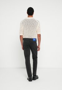 Valge lühikeste varrukatega kampsun avatud kudumismustriga, sobitatud tumedad hallid teksade ja mustade kingadega. Vöökohal sinine HUGO silt.