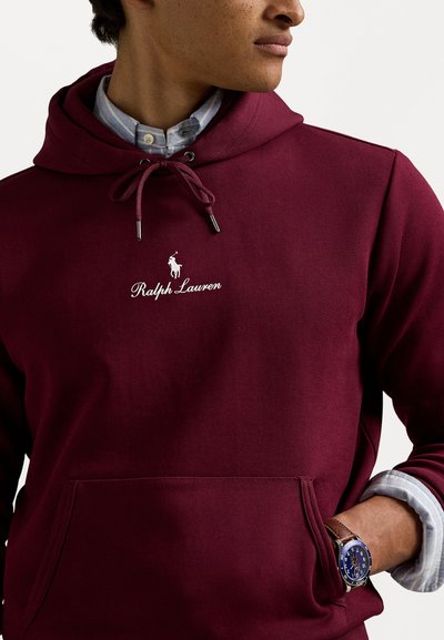 Moleton marrom feito de uma mistura de algodão, com um bolso frontal, cordões e um logotipo bordado em branco. O modelo usa uma camisa com colarinho por baixo.