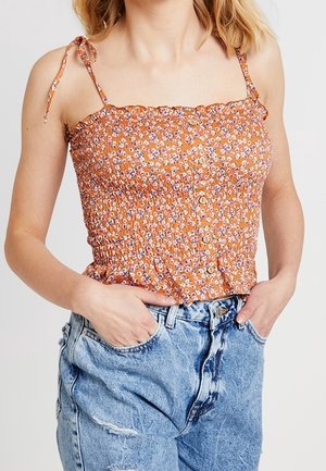 Femme portant un top orange fleuri smocké à fines bretelles avec des boutons, associé à un jean taille haute bleu clair déchiré.