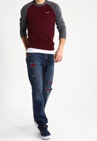 Uomo che indossa un maglione a maniche lunghe bordeaux e grigio, una maglietta bianca sotto, jeans blu strappati con toppe rosse e sneakers navy mentre cammina in avanti.