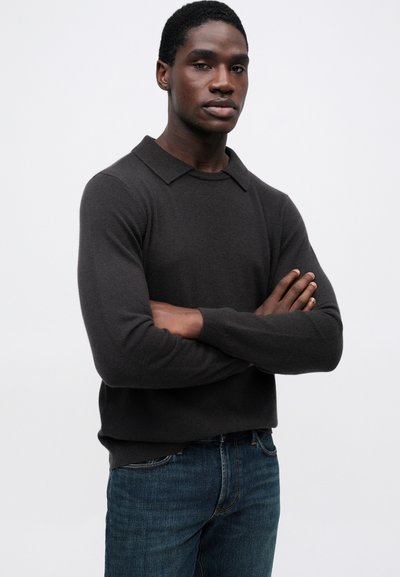 Fiatal férfi keresztbe tett karokkal, sötét hosszú ujjú pulóverben és kék farmerben áll egy sima fehér háttér előtt.