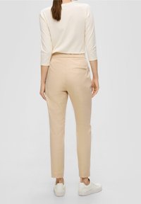 Frau mit braunen Haaren, die beige Hosen, ein weißes Langarmshirt und weiße Turnschuhe trägt, steht mit dem Rücken zu einer einfarbigen Hintergrund.