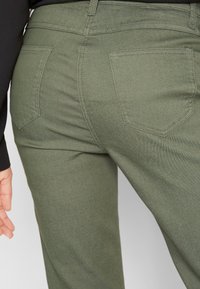 Pantalon vert foncé à texture lisse, avec deux poches arrière et des détails de coutures visibles. La ceinture est dotée de passants pour ceinture.