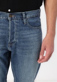 Niebieskie dżinsy z prostymi nogawkami, pojedynczym zapięciem na guzik i widocznymi detalami przeszycia. Tkanina ma wyblakły wygląd.