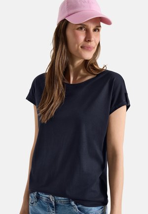 Femme aux longs cheveux bruns portant une casquette rose, un t-shirt bleu marine à manches courtes et un jean bleu, regardant légèrement sur le côté avec une expression neutre.