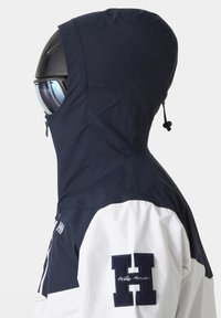 Námořnická a bílá větrovka s kapucí, zipem v přední části a vyšitým logem na rukávu. Materiál vypadá hladce a lehce.
