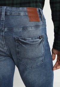 Blå denimjeans med en urtvättad finish, brun läderlogotyp på midjebandet och en bakficka med en vävd etikett.