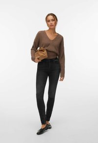 Femme debout portant un jean skinny noir, un pull côtelé marron à col en V, des ballerines noires, tenant une pochette en daim beige sur un fond blanc.