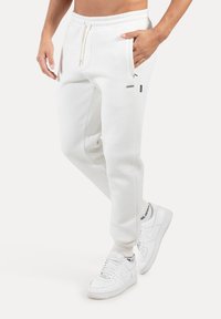 Pantalones deportivos de algodón blanco con cintura elástica y cordón ajustable. Presentan bolsillos laterales y un pequeño logo, combinados con zapatillas blancas.