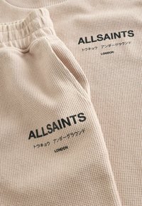 Beežika tekstuuriga kangast riided, millel on "ALLSAINTS" logo, Jaapani tähemärgid ning "LONDON" trükitud musta värviga vöö- ja rinnaosadel.