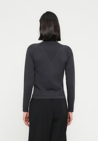 Femme aux cheveux bruns mi-longs portant un haut gris foncé cintré à manches longues et un pantalon noir, debout devant un fond blanc uni.