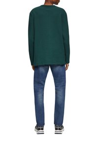 Groene langesleeve shirt met een ontspannen pasvorm, gecombineerd met blauwe jeans. De schoenen zijn zwart met witte accenten en hebben een sportief ontwerp.