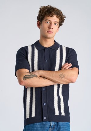 Jonge man met krullend haar, gekleed in een marineblauwe gebreide polo met witte strepen, staand met gekruiste armen en tattoos op de onderarmen toonend.