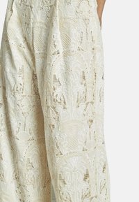 Pantaloni in pizzo con motivi intricati in bianco sporco, con una superficie texturizzata e ricami dettagliati su tutto il tessuto.
