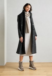 Manteau long noir avec un motif texturé, robe en maille grise, écharpe beige et bottes à lacets marron avec une semelle blanche, sur un fond neutre.
