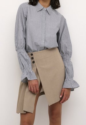 Vrouw draagt een zwart-wit gestreepte blouse met lange mouwen en ruches aan de manchetten, gecombineerd met een beige asymmetrische minirok met knoopdetails.