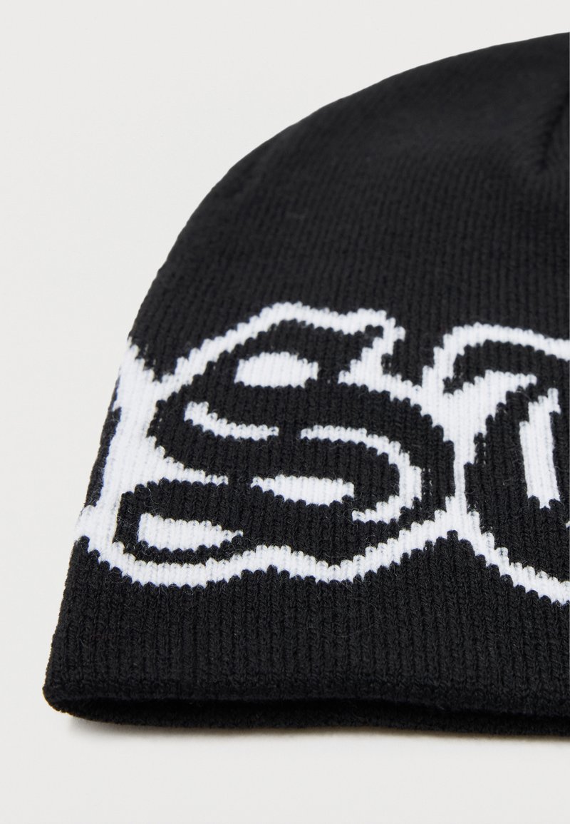 Beanie nero in maglia con design grafico bianco che mostra un testo stilizzato. Il materiale sembra morbido e strutturato, con una vestibilità aderente all'apertura.