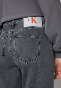 Jeans de mezclilla negra con cintura alta y cinco bolsillos, con un parche de cuero blanco que lleva el logo rojo "CK" en la parte posterior de la cinturilla.