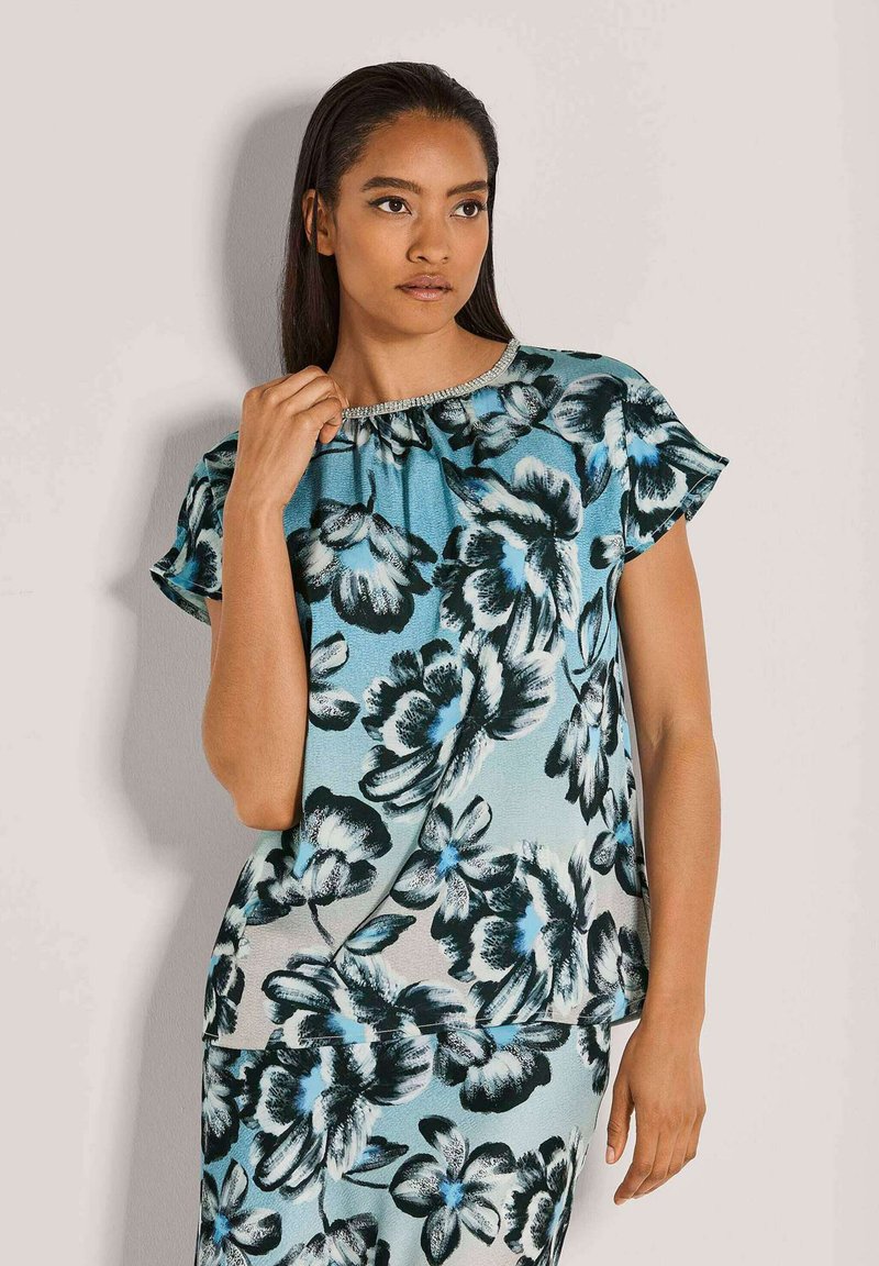 Blumenbluse in Blau und Schwarz mit kurzen Ärmeln, die einen gerafften Ausschnitt mit dezenten Verzierungen aufweist. Weicher, gemusterter Stoff.