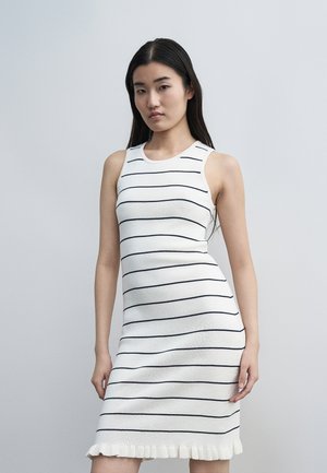 Γυναίκα με μακριά σκοτεινά μαλλιά φοράει αμάνικο λευκό φόρεμα με λεπτές μαύρες οριζόντιες ρίγες και κροσσί στο τελείωμα, στέκεται μπροστά σε απλό φωτεινό φόντο.