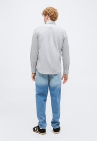 Homme aux cheveux bouclés debout de dos, portant une chemise à manches longues gris clair, un jean bleu délavé et des baskets noires avec des rayures blanches.