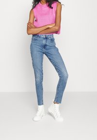 Top sin mangas de punto rosa con cuello alto, combinado con jeans ajustados azul claro y zapatillas blancas de caña alta.